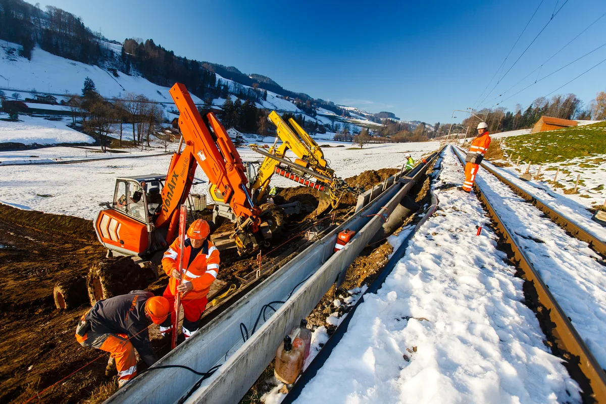 Bauarbeiter und Bagger an einem Gleisdamm am Arbeiten. 